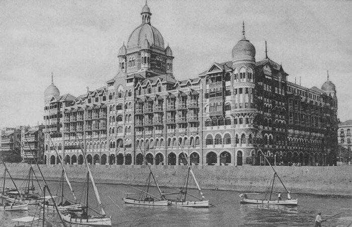 Historic black and white photograph of the Taj Mahal Palace hotel in Mumbai seen from the water, with sailboats in the foreground